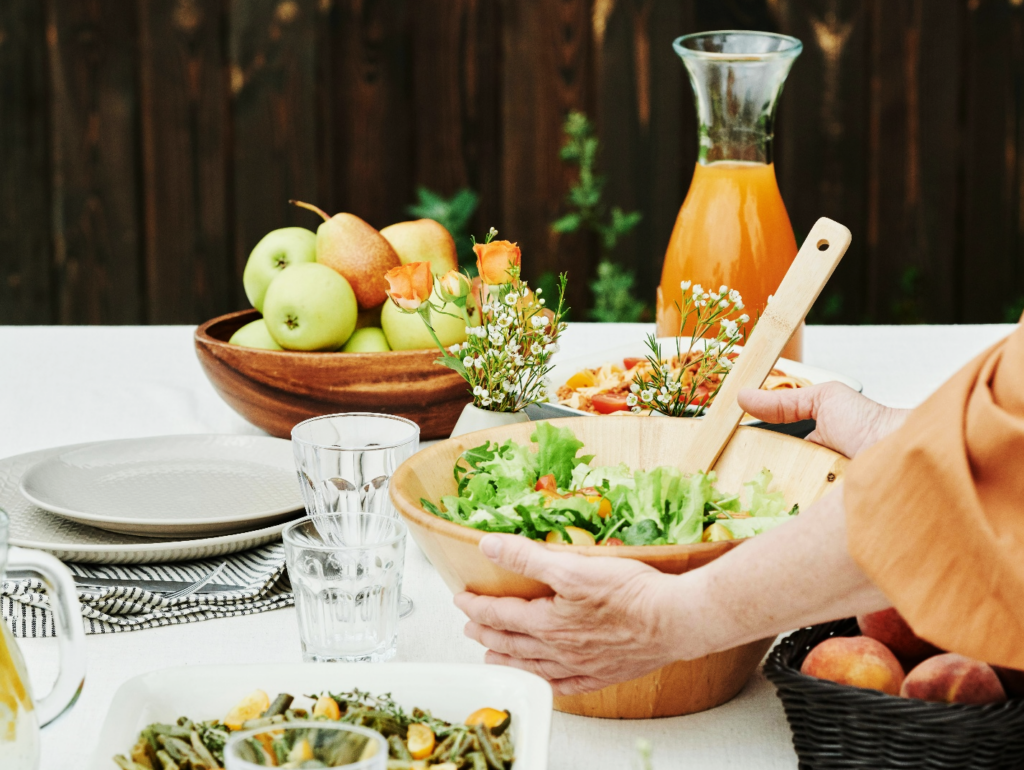 A dining table set for a meal, likely outdoors, with various dishes prepared and served. The table is covered with a white tablecloth and includes a salad, fruit, pasta or vegetables. bread, drinks, flowers, and tableware.