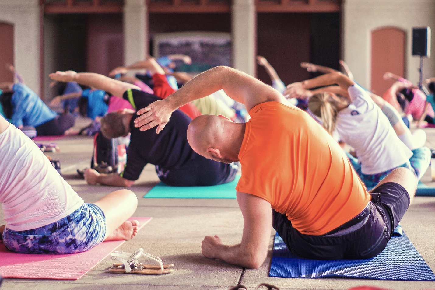 A group of people participating in a yoga class, seated on colorful mats in an open indoor space. The focus is on a bald man in an orange shirt performing a side stretch, with others in the background mirroring the same pose. A pair of sandals is visible on the ground near the mats.