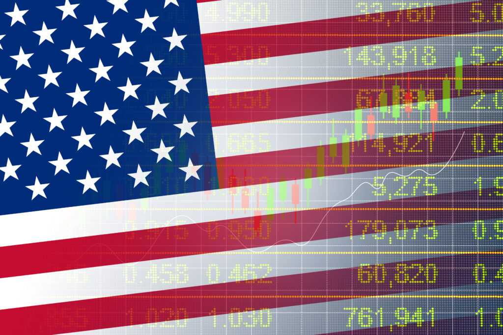 The American flag is overlaid on a background of stock market data, including green and yellow numbers and candlestick charts, symbolizing the intersection of U.S. economic policies and financial markets.