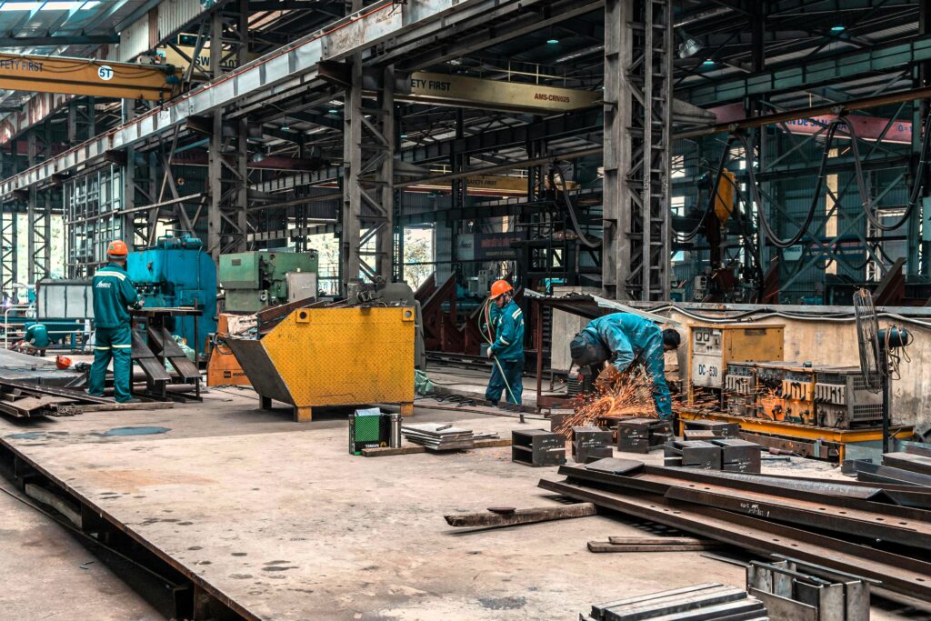 Workers in a large industrial factory wearing safety gear, with one operating machinery and another welding, producing sparks. The environment is filled with heavy equipment and steel structures.