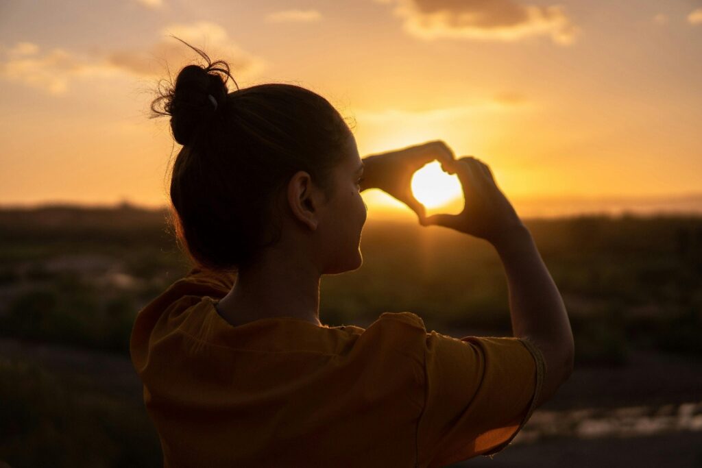 A person stands outdoors at sunset, forming a heart shape with their hands to frame the glowing sun, with a scenic landscape and soft clouds in the background.