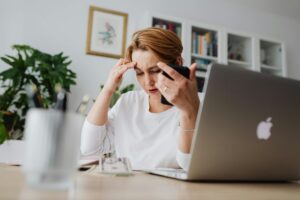 A woman sitting at a desk with a laptop, holding a phone to her ear and touching her forehead, looking worried as she deals with a possible bank account hacking.